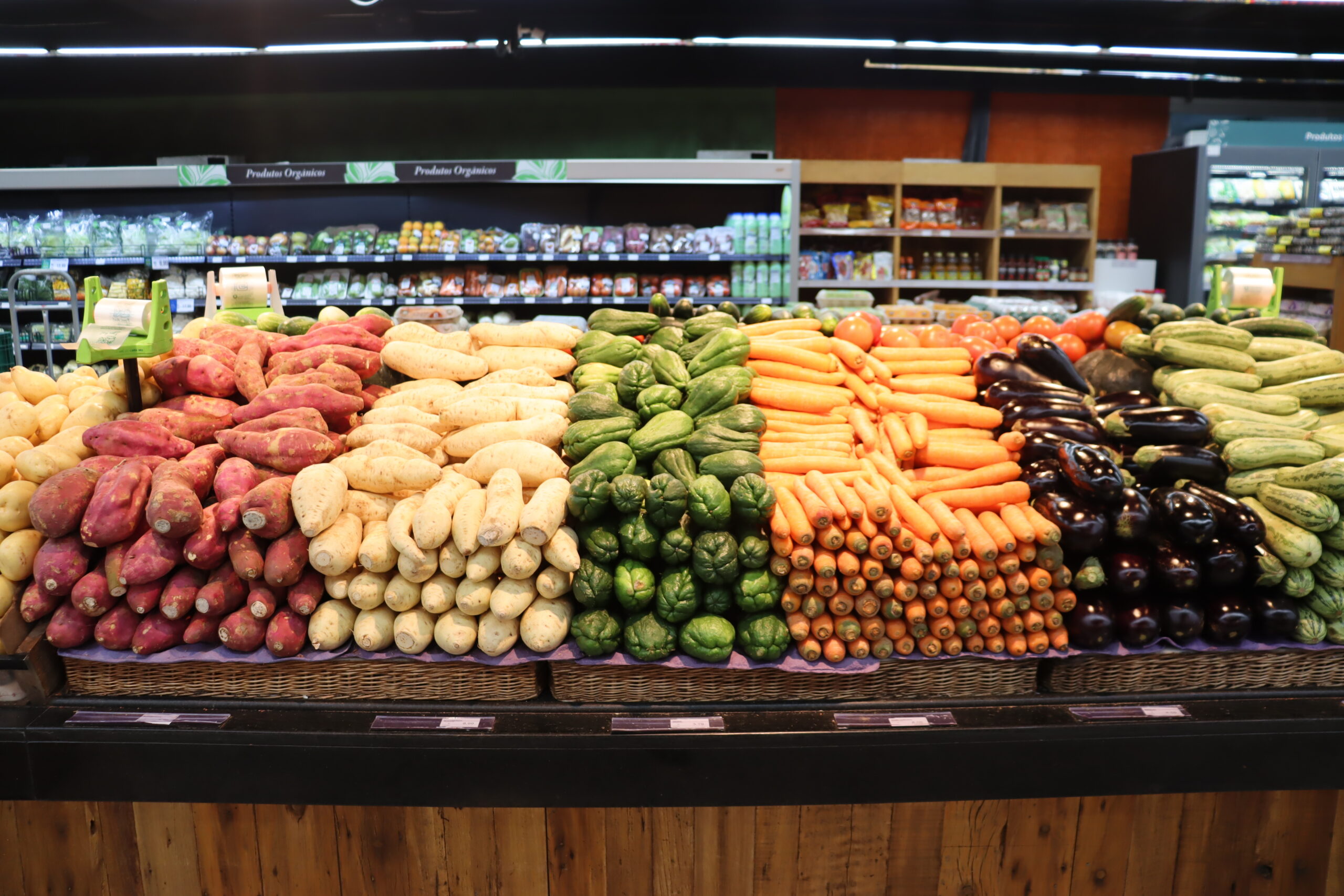 A imagem é composta por uma banca de legumes em um supermercado , os legumes dispostos são: batata doce, batata baroa, chuchu, cenoura, berinjela e abobrinha
