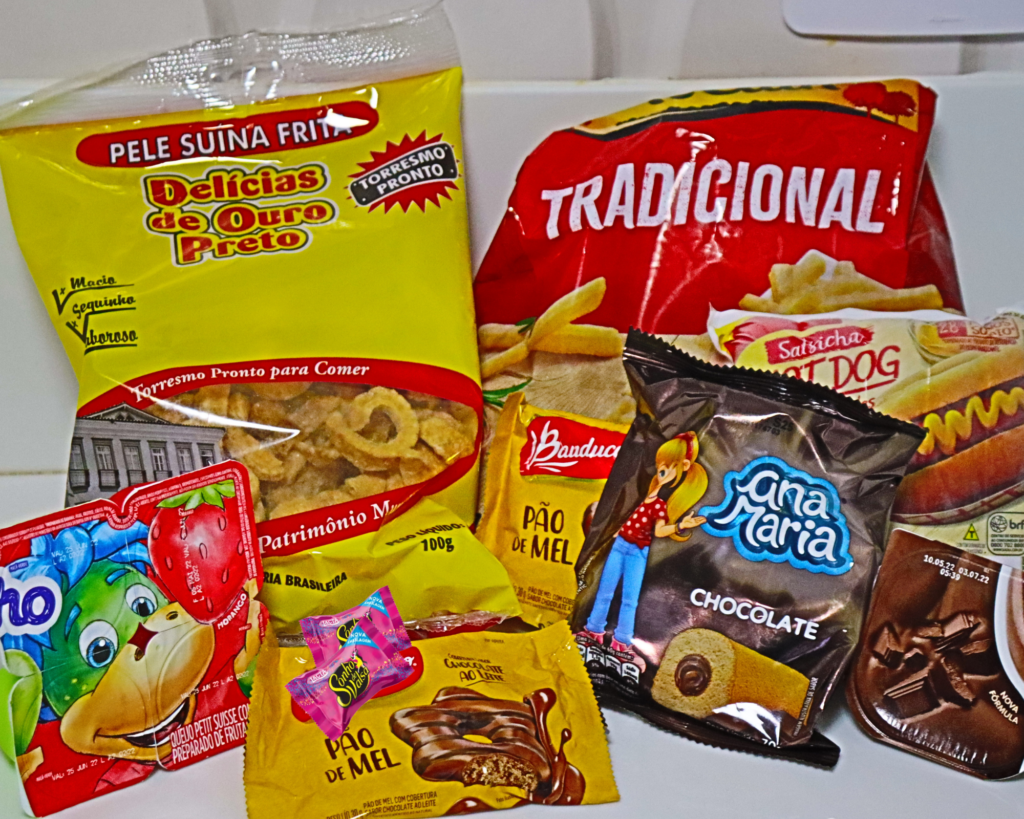 A imagem apresenta diversas embalagens de alimentos industrializados como: torresmo, bolinho, biscoitos, danoninho, danete, salsichas e batata frita