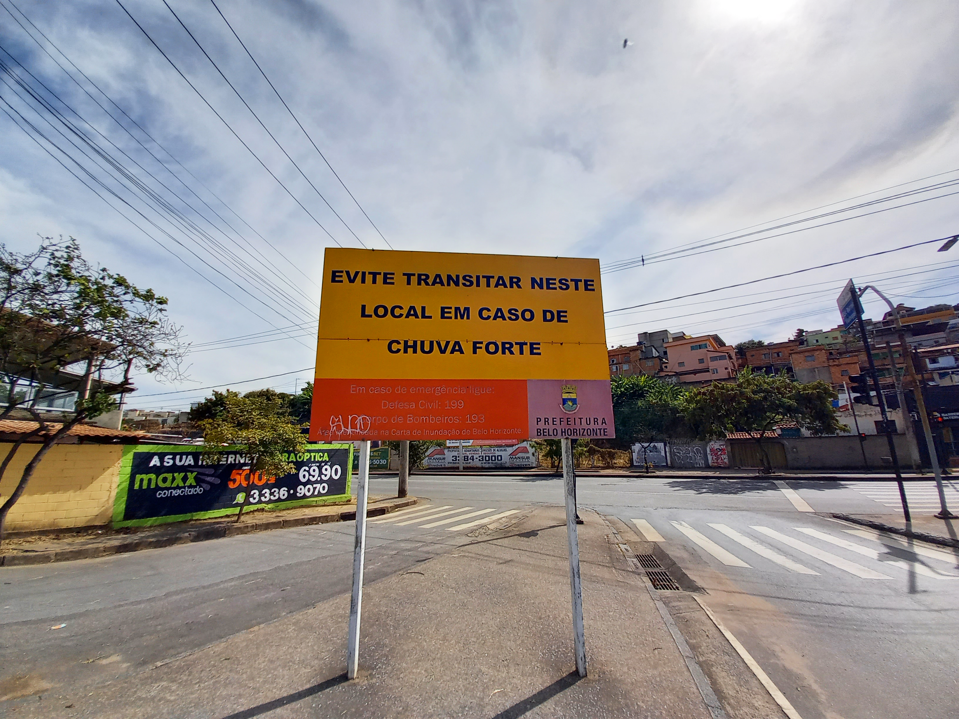 Foto diúrna da avenida Tereza Cristina. Em primeiro plano há uma placa amarela onde está escrito: "Evite transitar neste local em caso de chuva forte".