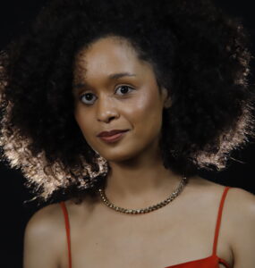 Jornalista Yasmin Santos. Mulher negra, jovem, com cabelo crespo volumoso, usa pouca maquiagem, um colar marrom e uma blusa vermelha.