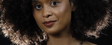 Jornalista Yasmin Santos. Mulher negra, jovem, com cabelo crespo volumoso, usa pouca maquiagem, um colar marrom e uma blusa vermelha.
