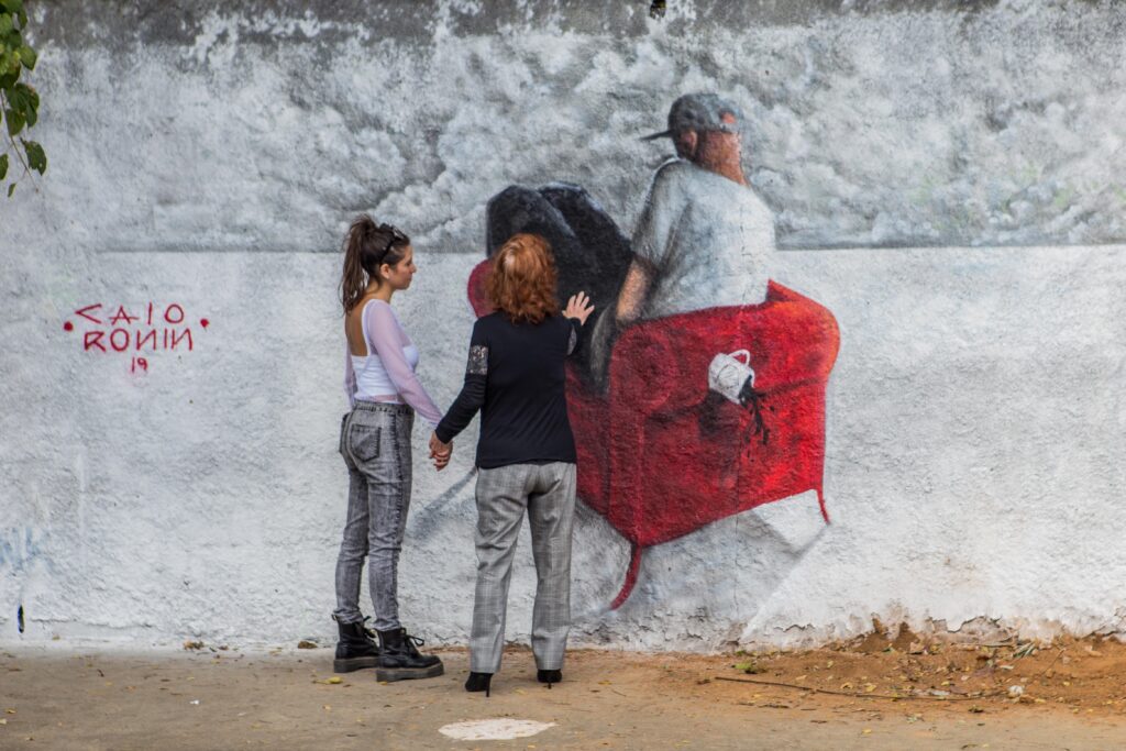 Visitantes interagem com painel feito para o TAU em 2019. Duas mulheres estão de mãos dadas, de costas. Uma delas encosta a mão na pintura, que é branca e vermelha. 