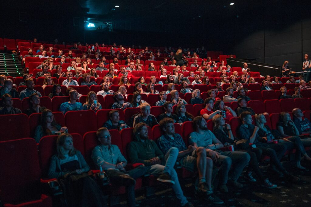 Sala de cinema com cadeiras vermelhas, cheia de pessoas focadas no que está sendo passado na tela durante uma sessão de cinema.