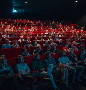 Sala de cinema com cadeiras vermelhas, cheia de pessoas focadas no que está sendo passado na tela durante uma sessão de cinema.