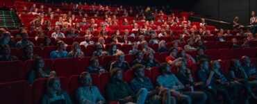 Sala de cinema com cadeiras vermelhas, cheia de pessoas focadas no que está sendo passado na tela durante uma sessão de cinema.