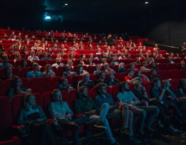 Sala de cinema com cadeiras vermelhas, cheia de pessoas focadas no que está sendo passado na tela durante uma sessão de cinema.