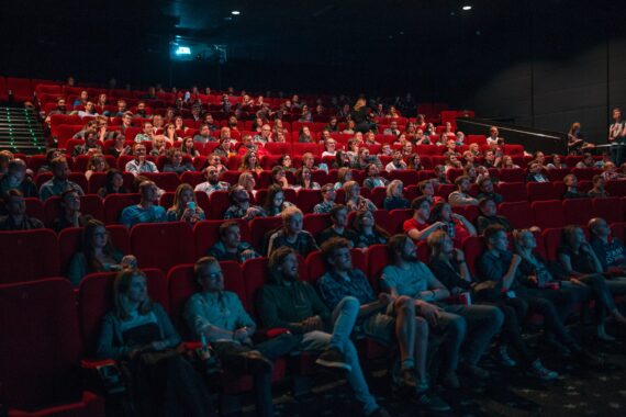 Sala de cinema com cadeiras vermelhas, cheia de pessoas focadas no que está sendo passado na tela durante uma sessão de cinema.