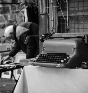 Imagem em preto e branco em que há uma máquina de escrever em primeiro plano e, em segundo plano, um senhor abaixado mexendo em algo.