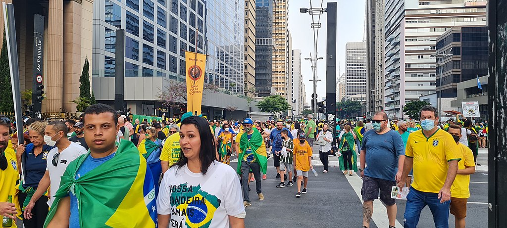 Uma multidão pessoas vestem camisas com as cores verde, amarelo e azul enquanto caminham pela Avenida Paulista. Alguns possuem a bandeira do Brasil amarrada em volta do pescoço.