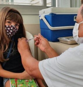 Foto que contém uma menina branca do cabelo cumprido e claro com um senhor aplicando a vacina em seu braço. Ao fundo há uma parede clara e um recipiente azul e branco para guardar as vacinas.