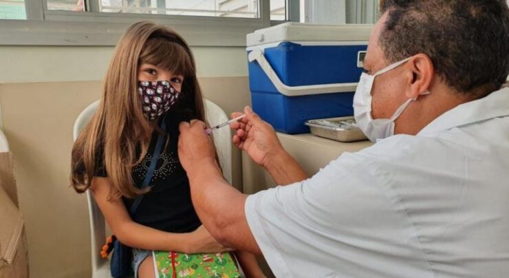 Foto que contém uma menina branca do cabelo cumprido e claro com um senhor aplicando a vacina em seu braço. Ao fundo há uma parede clara e um recipiente azul e branco para guardar as vacinas.