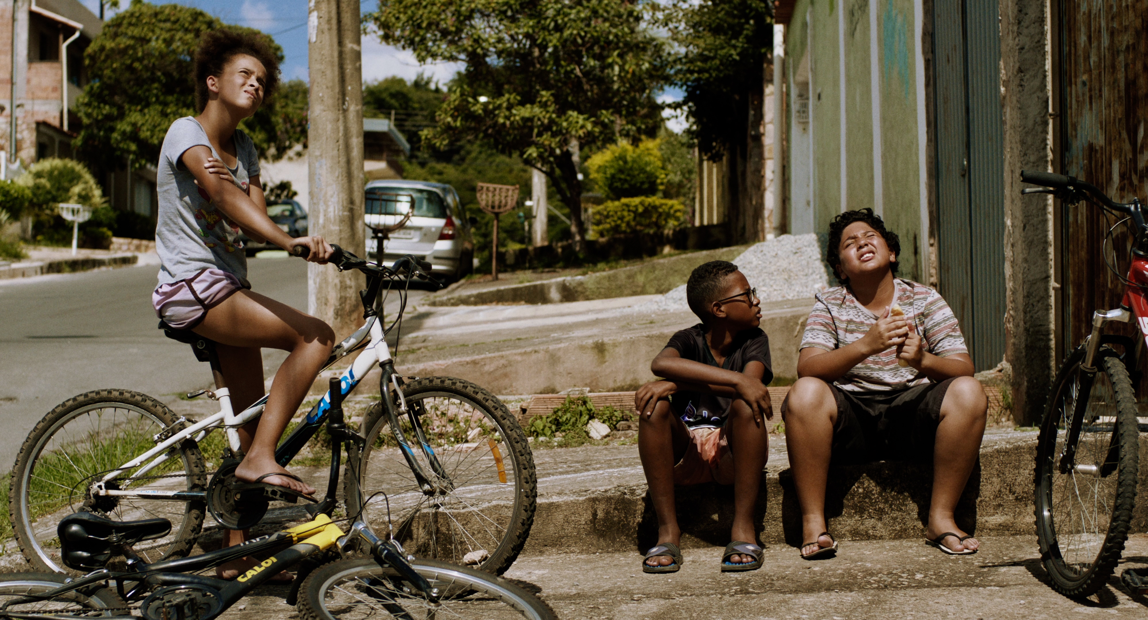 foto de dois meninos sentados no meio fio e uma menina sentada na bicicleta 