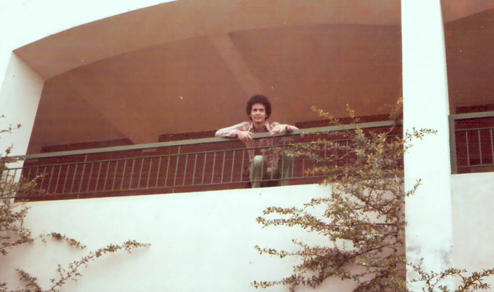 Foto de arquivo pessoal mostra o jovem advogado Gabriel Pimenta em uma varanda de grade verde sob estrutura de concreto branco, no segundo andar de um dos edifícios da Universidade Federal de Juiz de Fora. Ele veste blusa xadrez clara e calça. Atrás dele é possível ver uma parede de tijolinhos. .