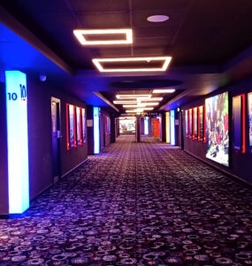 Um corredor de cinema é iluminado pelas luzes do teto e por cartazes luminosos dos filmes em cartaz na paredes laterais