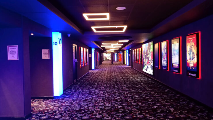 Um corredor de cinema é iluminado pelas luzes do teto e por cartazes luminosos dos filmes em cartaz na paredes laterais