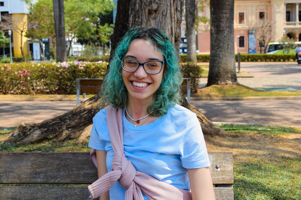 Menina de óculos, cabelos verdes e sorridente, sentada em um banco de uma praça em um ambiente externo