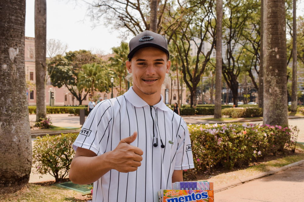 Foto de um menino vendedor de balas segurando uma caixa de "mentos" sorrindo enquanto faz um "joinha" 