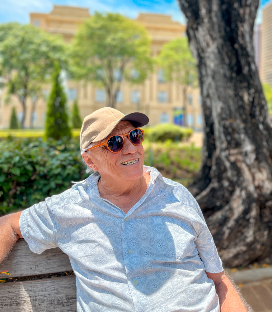 Foto de um senhor idoso sentado em um banco da praça, ele usa um óculos escuro colorido e um boné enquanto sorri olhando para o horizonte 