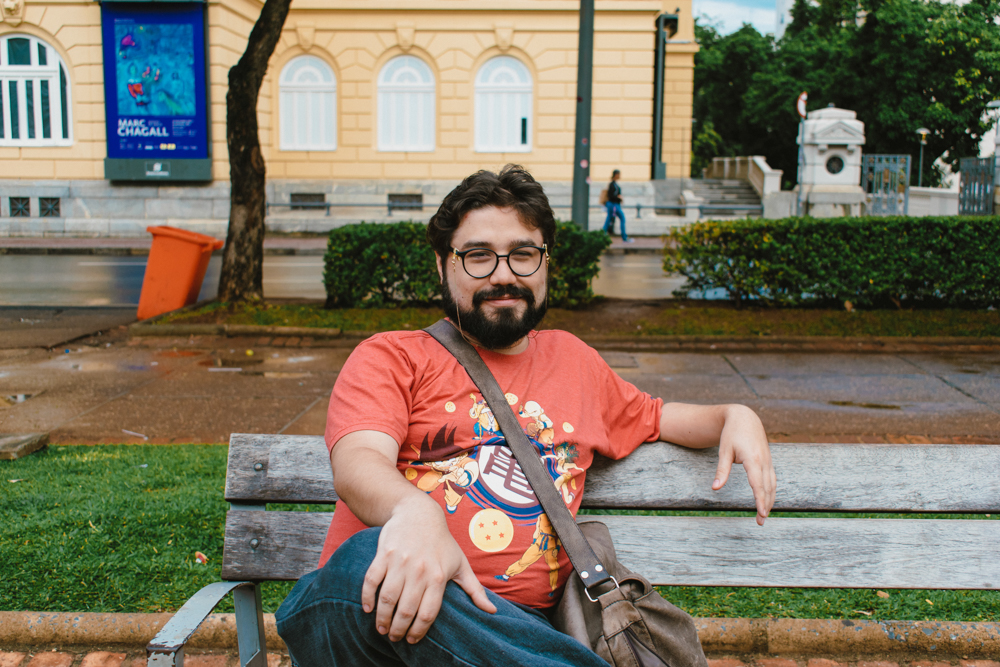 Foto de um homem barbudo, usando óculos. Ele está cruzando as pernas enquanto senta no banco da praça e sorri timidamente