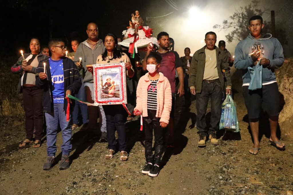 Moradores de Bento Rodrigues visitam as ruínas da cidade durante a noite para festa religiosa. Pessoas se agrupam, carregam imagens de Nossa Senhora e velas.