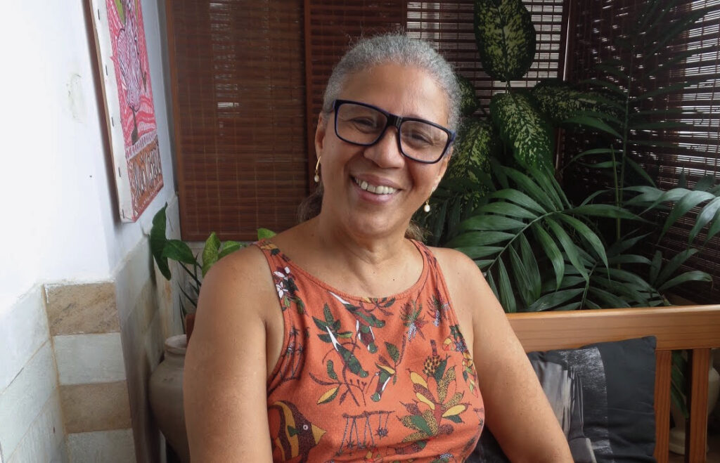 Ao centro da foto, uma mulher negra que está sorrindo. Ela tem cabelos brancos e veste uma camiseta regata de cor laranja com estampas de flores e plantas. Ela usa óculos e brincos. A mulher está sentada em uma cadeira de madeira com almofadas pretas. Atrás dela, ao fundo da foto, há cortinas no tom marrom e vários vasos de plantas no chão, além de um quadro de pintura na parede.