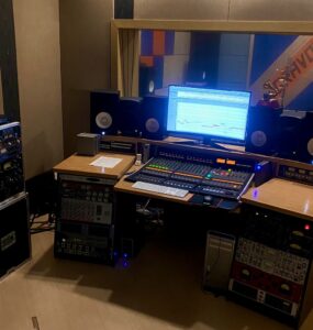Foto do estúdio principal do Estúdio Gravomix. Em primeiro plano, Mesa de som, computador com a tela ligada. Em segundo plano, estúdio de captação de áudio.