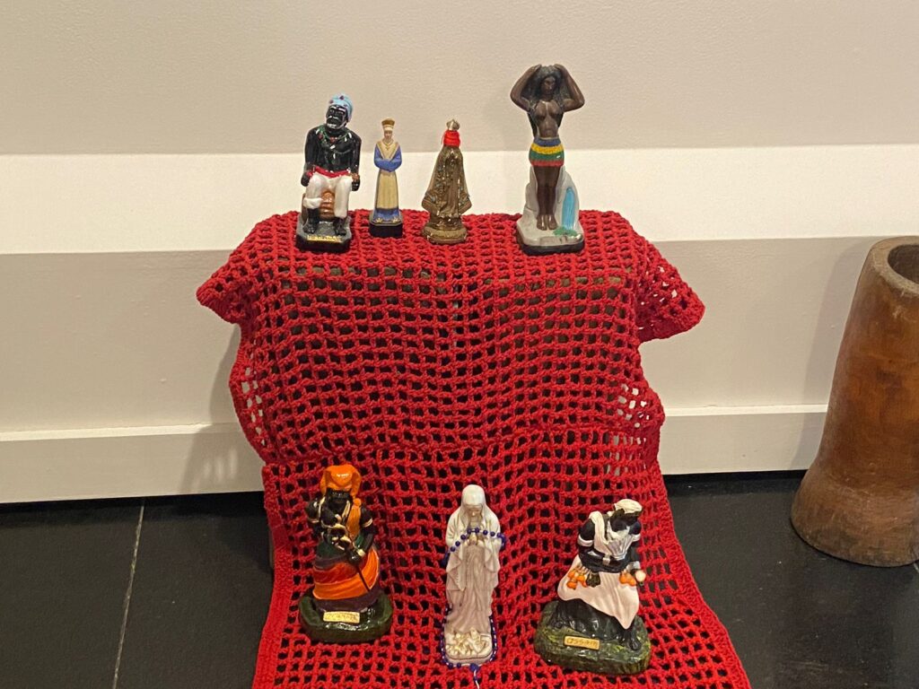 Altar coberto com trama de crochê vermelho. Sobre ele há diversas imagens de objetos sagrados para a Umbanda 