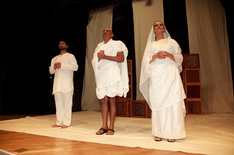A imagem mostra um palco de teatro em tons bege e marrom, com chão de madeira. No centro do palco estão os três atores, entre eles a atriz Patricia Naves (à direita de quem vê), vestidos com túnicas brancas.
