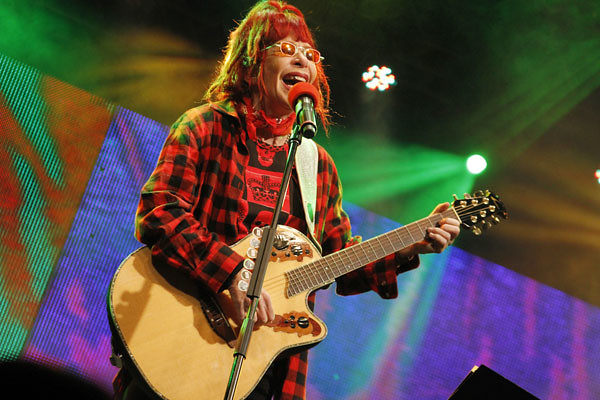 Cantora Rita Lee com seus cabelos e roupas vermelhas, toca violão e canta em frente ao microfone com luzes coloridas ao fundo.