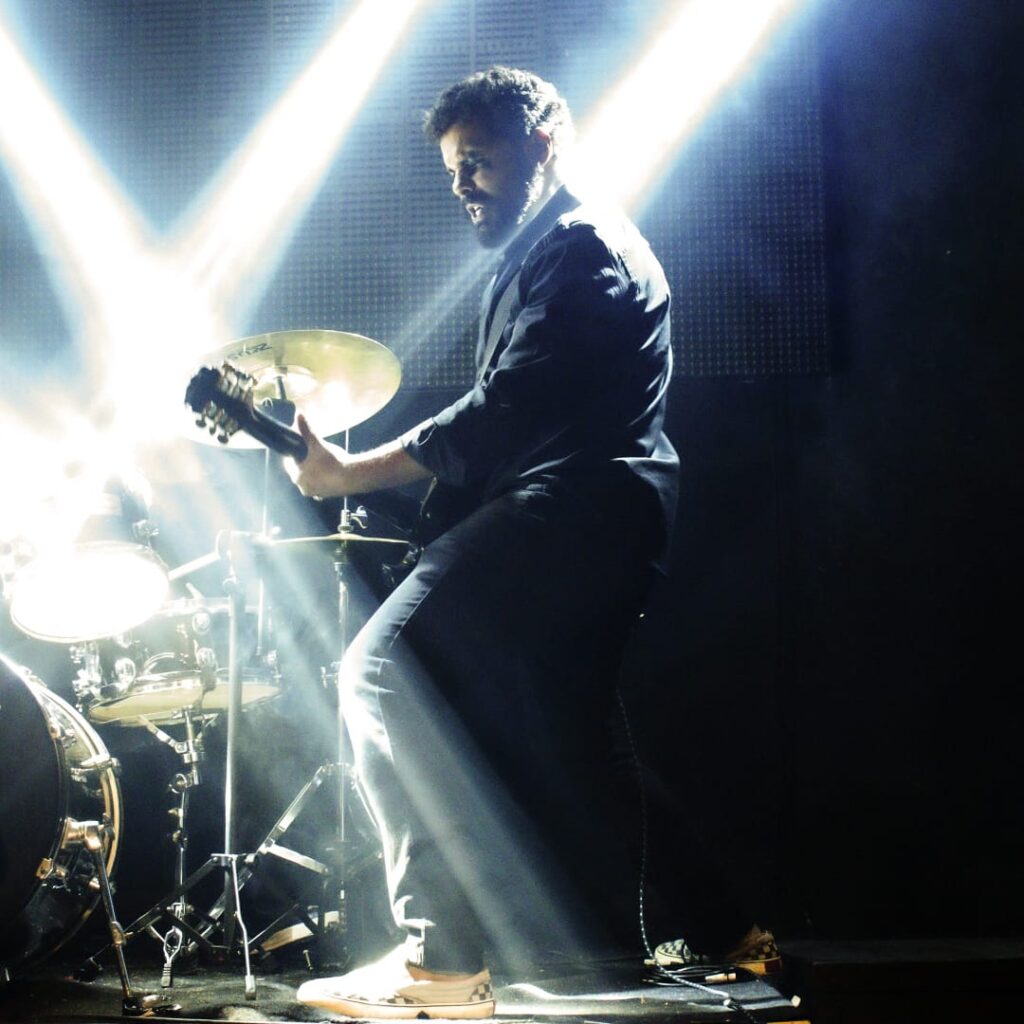 Cantor Rubah se encontra ao centro tocando guitarra ao lado de uma bateria. Duas faixas de luz vindas de cima de Rubah iluminam ele, uma em direção a guitarra e outra em sua nuca.