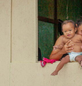 Mãe venezuelana beija costas de sua bebê ao lado de outra filha. Todas estão olhando fixamente para a câmera em frente à janela