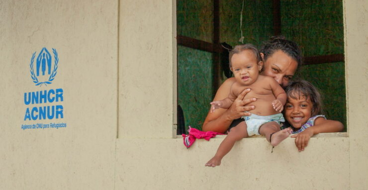 Mãe venezuelana beija costas de sua bebê ao lado de outra filha. Todas estão olhando fixamente para a câmera em frente à janela