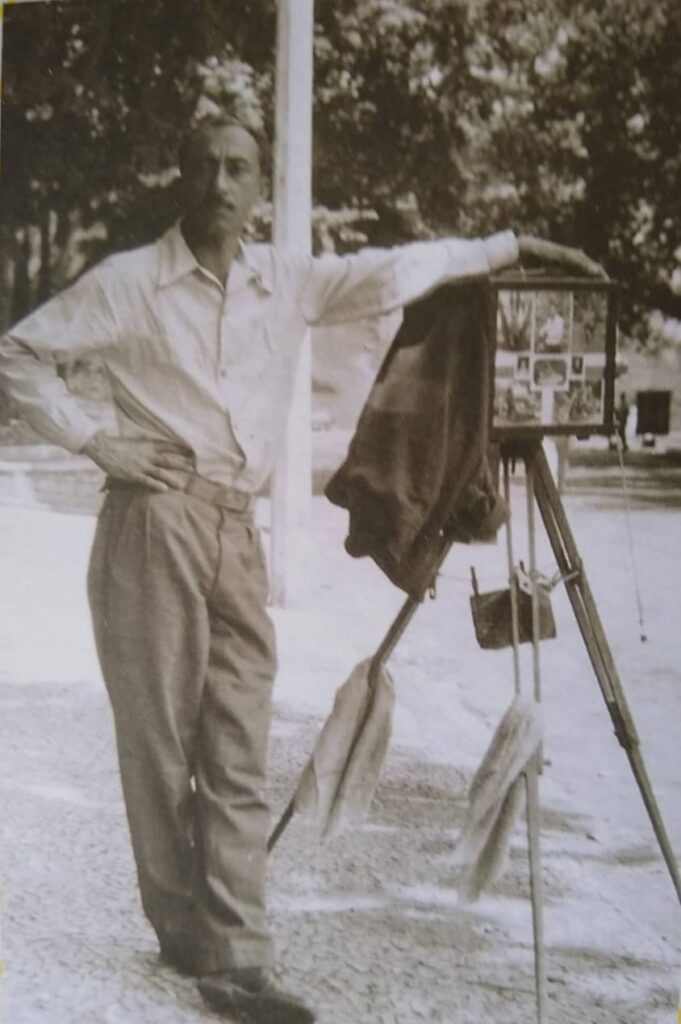 Descrição ALT: Foto em preto e branco de um homem adulto em pé, que veste uma camisa clara de botões e uma calça escura de alfaiataria. Ele tem cabelo curto, bigode e uma expressão séria no rosto. Está com uma perna à frente da outra, uma mão na cintura e outra apoiada em uma máquina caixote. Ele está no Parque Municipal de Belo Horizonte, com árvores ao fundo.