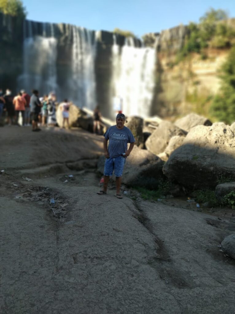 Descrição de imagem: Victor Hugo no centro da imagem vestindo uma camiseta cinza e bermuda jeans, em  frente à uma cachoeira.