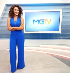 Aline Aguiar, jornalista negra, está vestindo um macacão azul. Ela está de pé em frente a uma tv, que reproduz o logo do MGTV.
