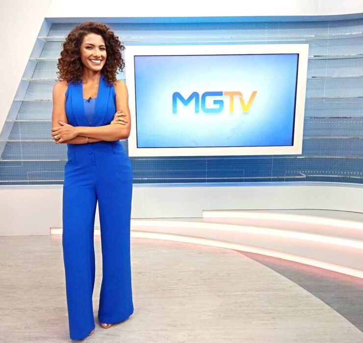 Aline Aguiar, jornalista negra, está vestindo um macacão azul. Ela está de pé em frente a uma tv, que reproduz o logo do MGTV.