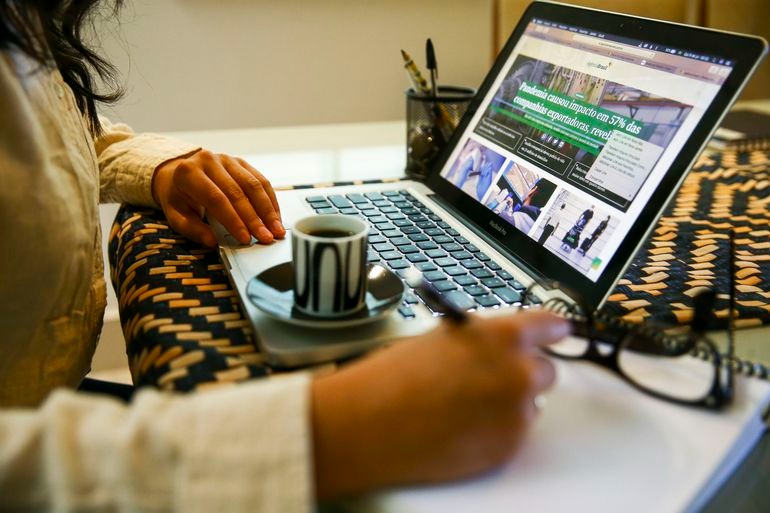 Descrição de imagem: mulher sentada, tomando café, mexendo no computador e anotando em um caderno.