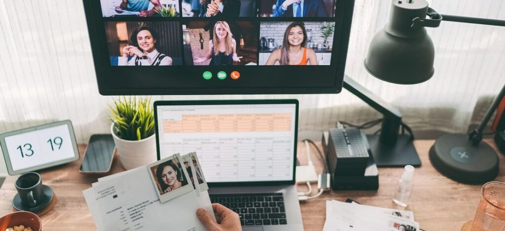 Descrição de imagem: ambiente de escritório, com computador aberto em uma tabela, uma tela a frente com pessoas na videochamada.