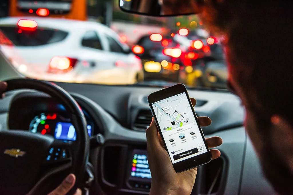 Descrição de imagem: homem dentro do carro e parado no congestionamento segurando o celular com o aplicativo do uber aberto. 