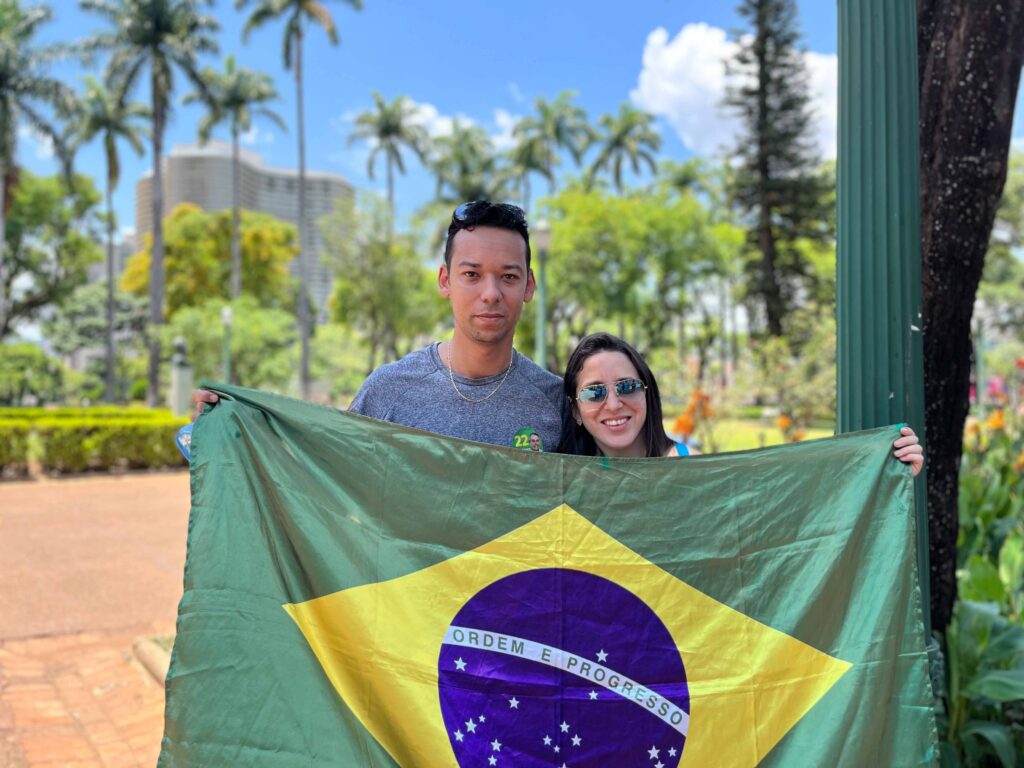 Foto de um casal composto por um homem pardo e uma mulher branca estendendo uma bandeira do Brasil na praça