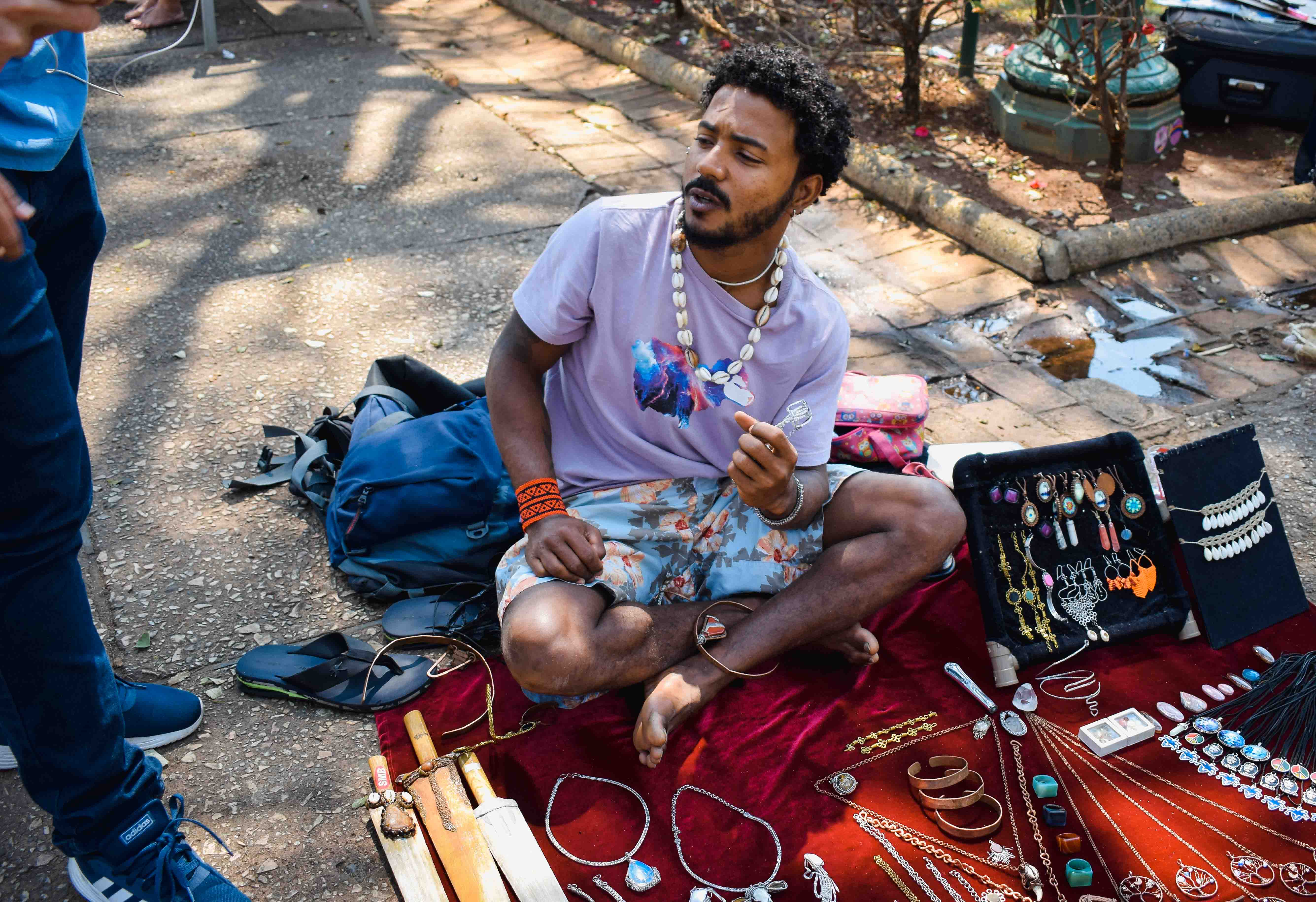 Foto de um vendedor de arte sentado no chão da praça exibindo seus artesanatos, ele usa um colar de conchas enquanto é entrevistado