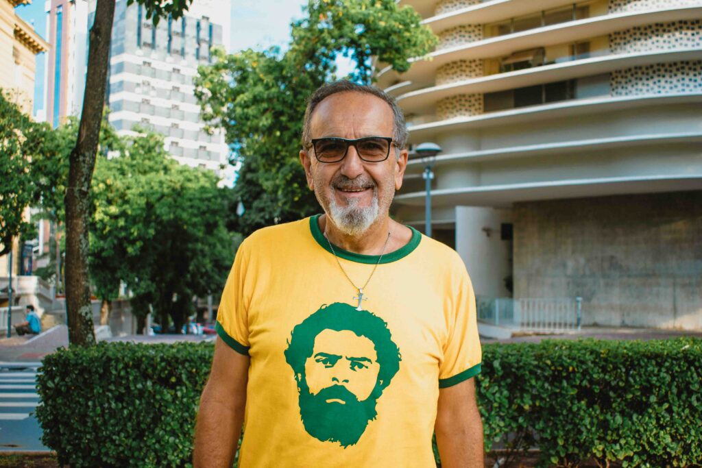 Foto de um homem sorridente de cabelos brancos vestido com uma camisa verde e amarela com o rosto de Lula estampado.