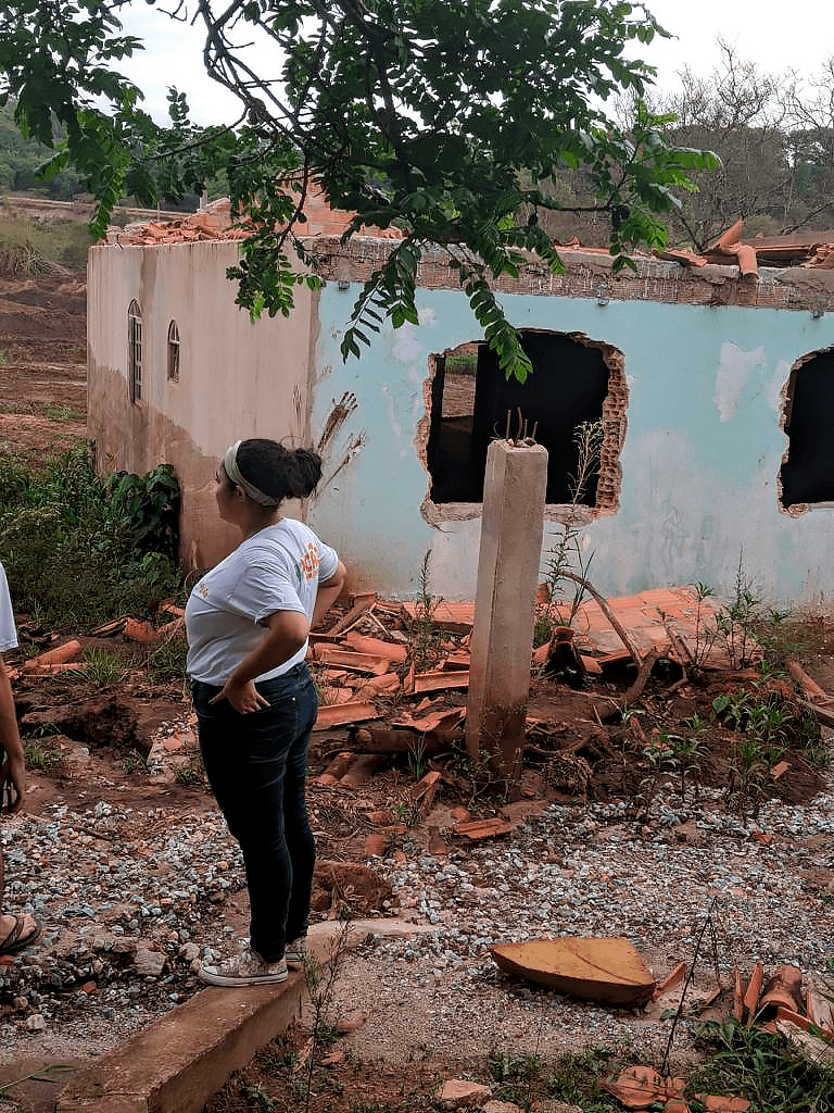 Imagem de casa destruída pela barragem, com mulher voluntária em primeiro plano, que está de cabelo preso e blusa da ONG, braços apoiados na cintura e virada de lado.