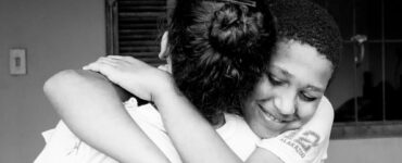 Imagem em preto e branco de duas pessoas se abraçando, uma mulher de costas e um jovem com expressão feliz de frente.