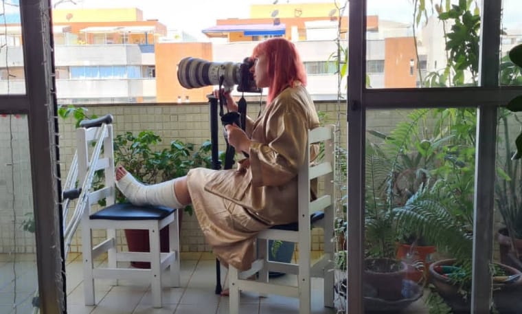 Descrição alt: Gabriela Biló, de cabelos rosa claro e roupão bege,  está sentada em uma cadeira branca e azul em uma varanda rodeada de prédios e alguns vasos de plantas, a fotógrafa repousa sua perna engessada sobre uma cadeira semelhante, enquanto faz registros de rua com o auxílio de um tripé e uma teleobjetiva (lente fotografia para grande alcance)
