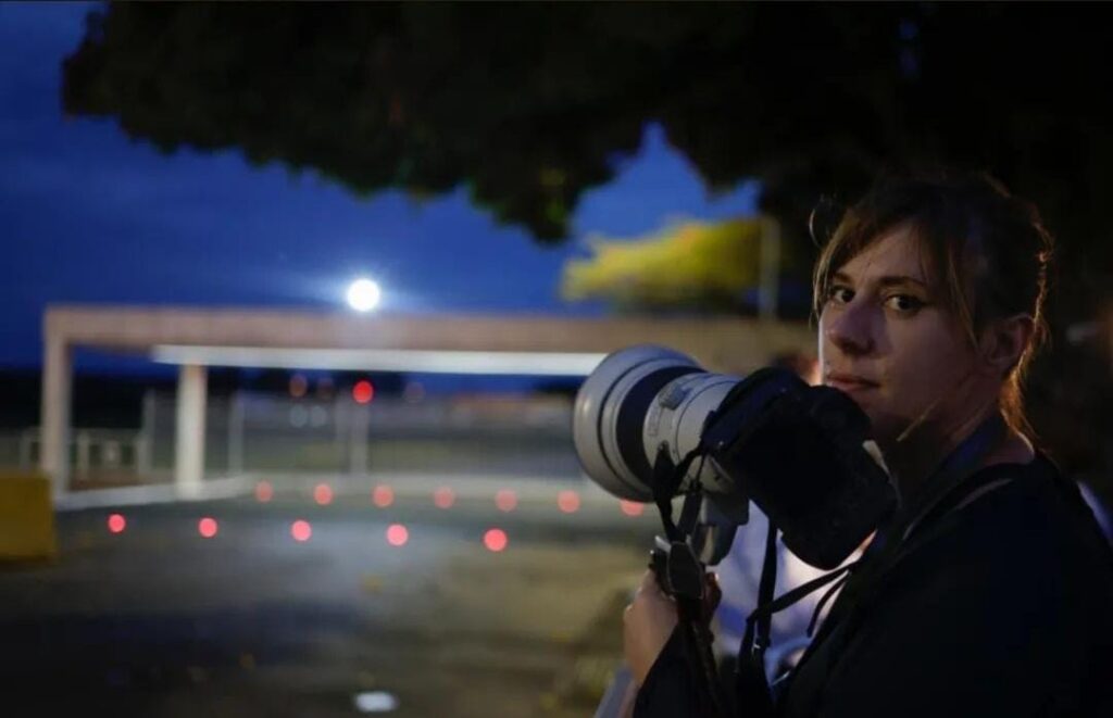 Gabriela Biló de frente ao Palácio do Planalto ao entardecer, a fotógrafa segura um teleobjetiva dentro do "cercadinho" reservado para a equipe de imprensa, a câmera próxima ao queixo e o recorte de luz lateral realçam a aparência seria e cabelos presos da fotógrafa