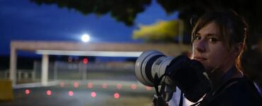 Gabriela Biló de frente ao Palácio do Planalto ao entardecer, a fotógrafa segura um teleobjetiva dentro do "cercadinho" reservado para a equipe de imprensa, a câmera próxima ao queixo e o recorte de luz lateral realçam a aparência seria e cabelos presos da fotógrafa