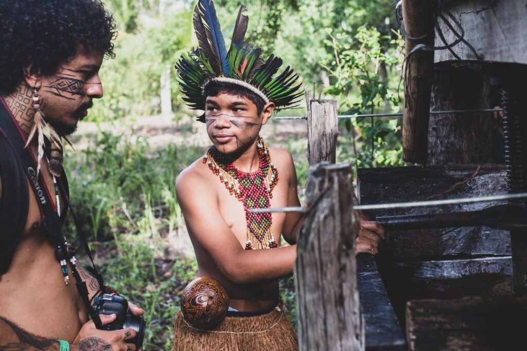 Emanuel Kaauara está localizado à esquerda da imagem com uma câmera fotográfica em mãos observando atentamente um indígena Pataxó que lhe explica um oficio.