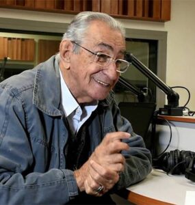 Ricardo Parreiras, cabelos grisalhos, sorridente .Ele usa óculos e jaqueta. Está sentado em frente a um microfone.