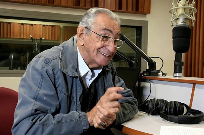 Ricardo Parreiras, cabelos grisalhos, sorridente .Ele usa óculos e jaqueta. Está sentado em frente a um microfone.